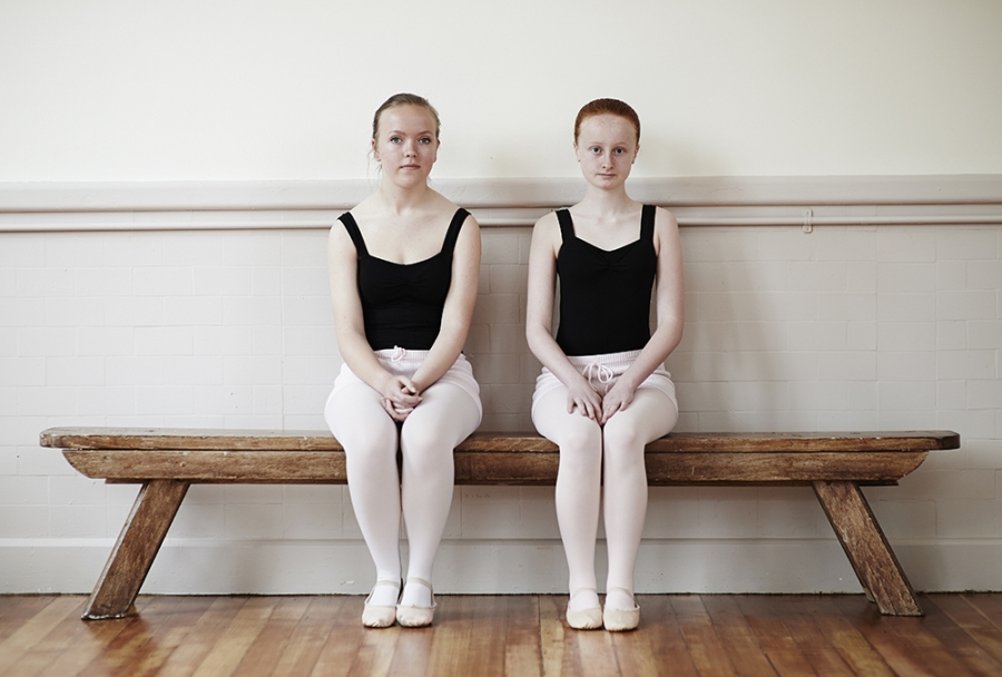Two female ballet pupils on bench looking to camera.